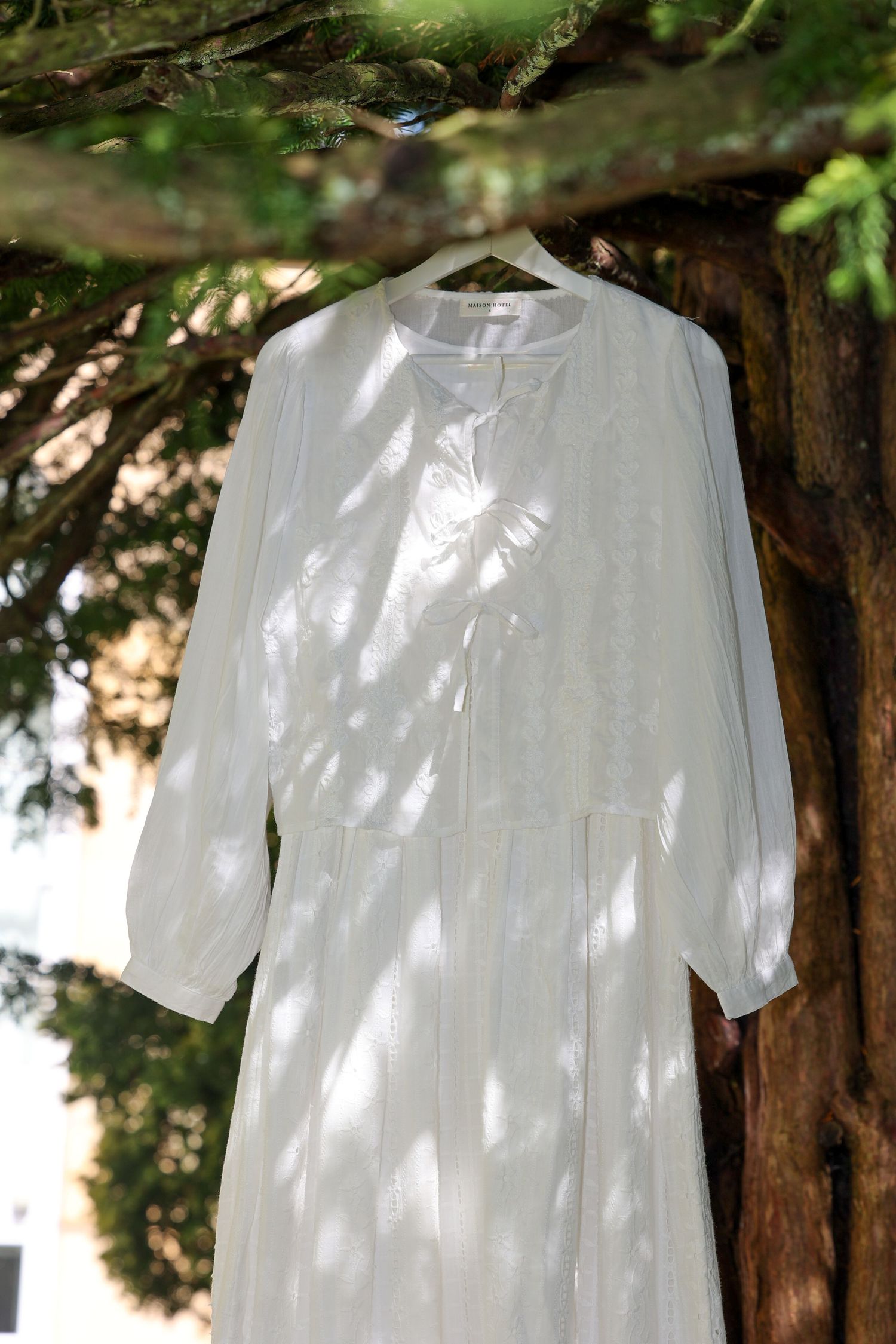 White long-sleeved linen dress hanging on a tree branch with dappled sunlight casting shadows through leaves.