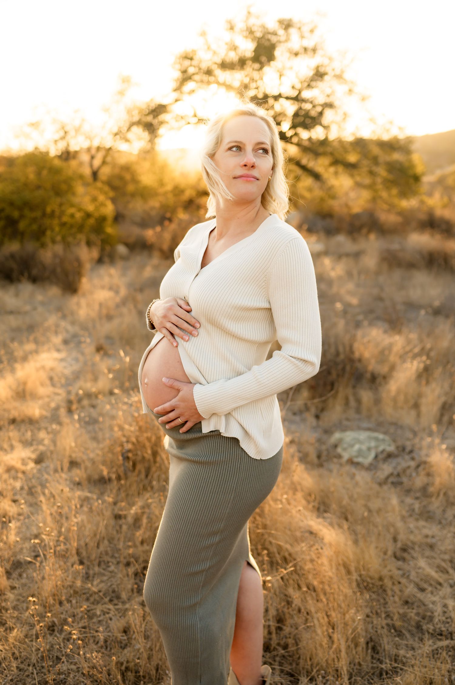 woman stands in golden sunlight holding bare pregnant belly during her Ojai babymoon maternity photoshoot, Ventura photographer