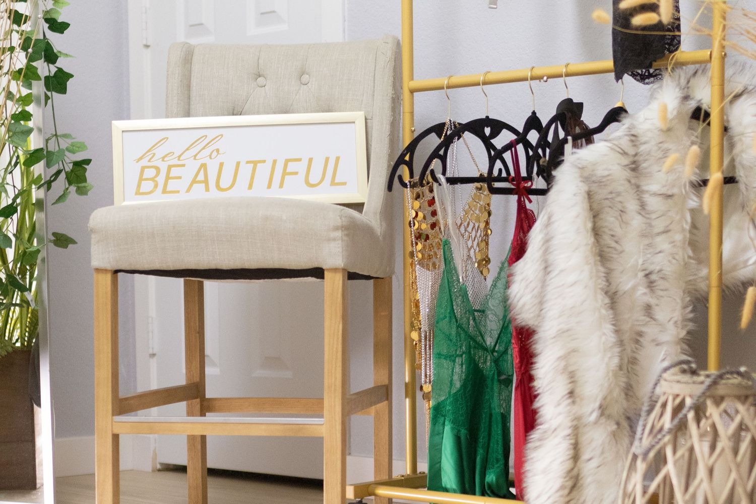 Beige upholstered barstool with 'Hello Beautiful' pillow next to gold clothing rack with dresses and fur coat.