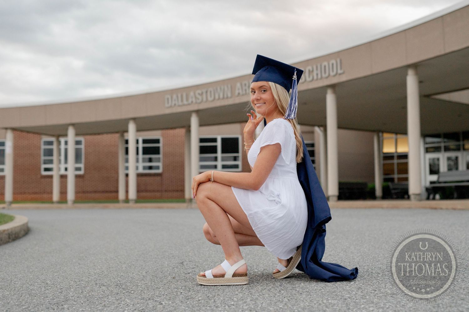 Graduate in white dress and blue cap and gown poses outside Dallastown Area High School on graduation day.