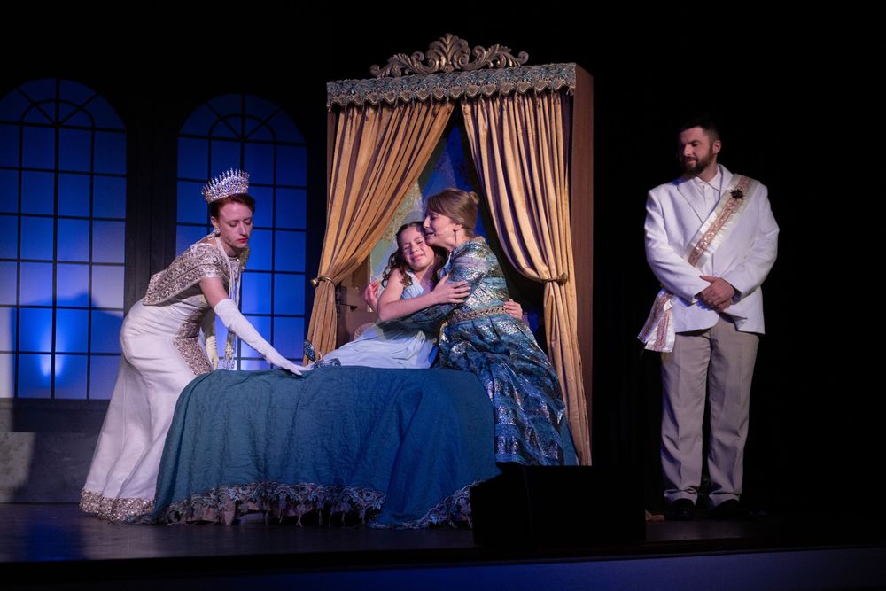 Theater performers act out a dramatic bedroom scene on stage with period costumes and ornate set design.