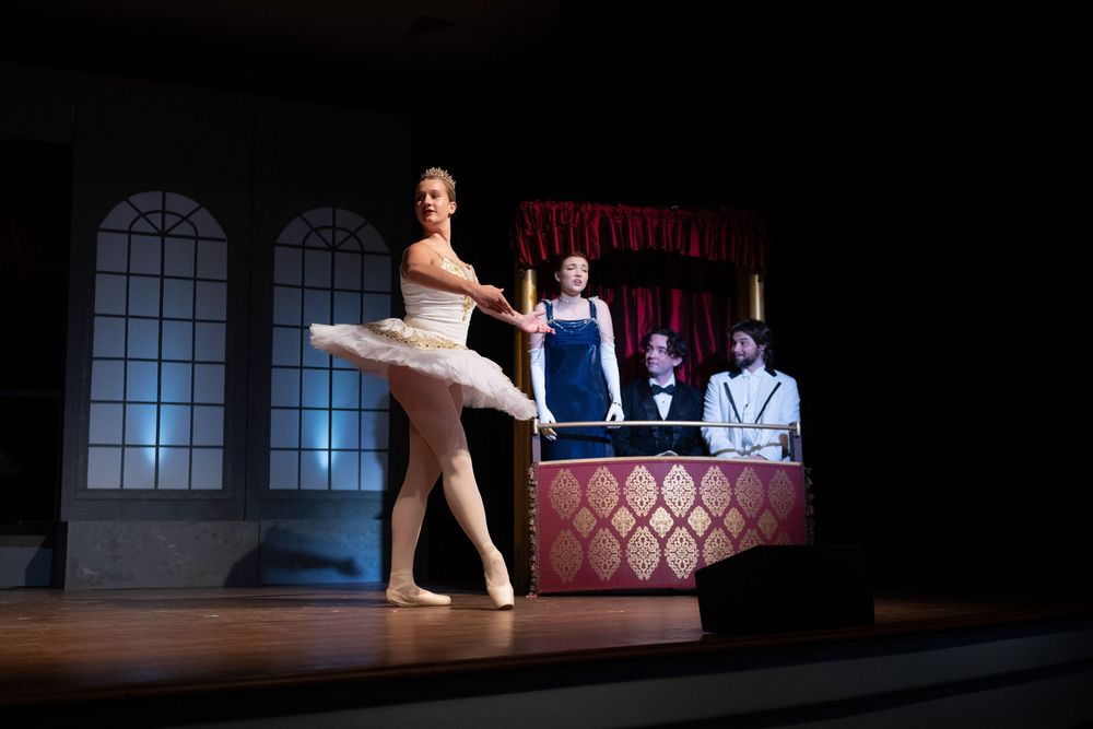 A ballerina in white tutu performs onstage in front of tall windows while musicians play at a raised platform.