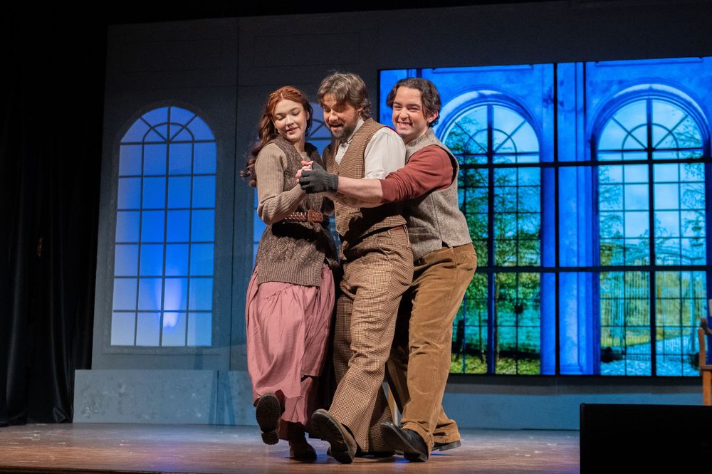 Three performers act together in a theatrical scene on stage with blue-lit arched windows in the background.