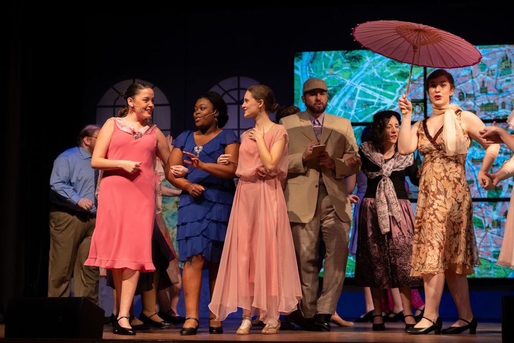 Cast members perform on stage in colorful dresses and costumes during a musical theater production.