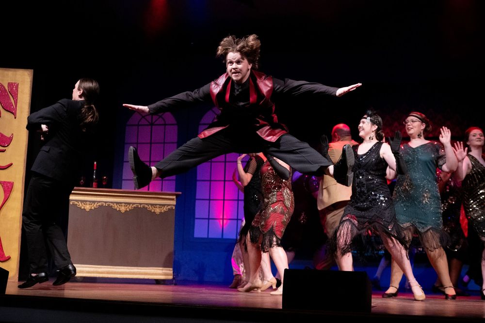 Performers on stage during a theatrical production with colorful lighting, featuring a person leaping dramatically over a prop.