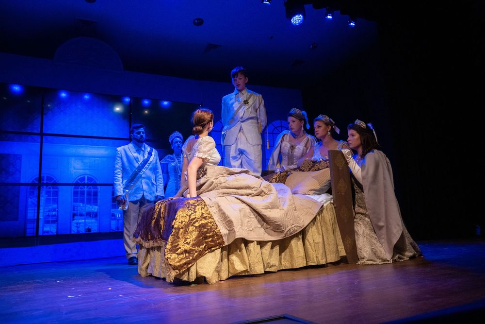 Actors perform in period costumes during a dramatic stage play scene with blue lighting and elegant Victorian-era dress.
