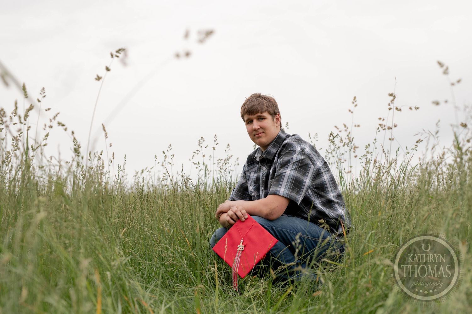 Person in plaid shirt sits in tall meadow grass surrounded by wildflowers on an overcast day.