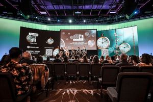 Audience members seated in rows watching a panel discussion on stage with digital screens displaying content in background.