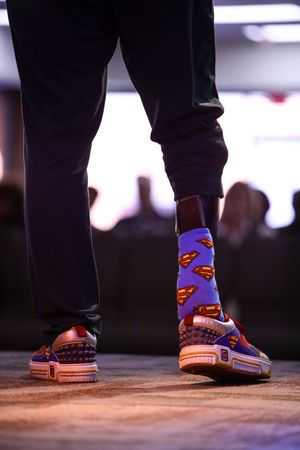 Person wearing one playful Superman print sock with black pants and casual sneakers standing on wooden stage.