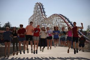 Group of excited people jumping in unison with a large wooden roller coaster structure visible in the background.
