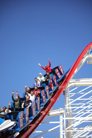 Thrill seekers raise their arms while riding a steep red and white roller coaster track against bright blue sky.