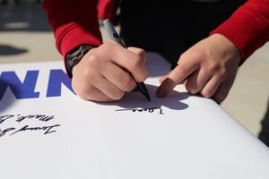 Close-up view of hands signing an autograph on a white surface with blue text visible in the background.