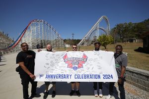 Small group holds up banner celebrating rollercoaster's first anniversary with towering red and white coaster track visible behind.