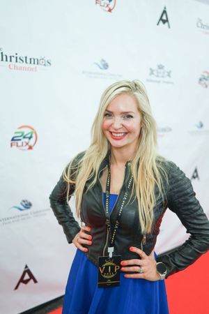 Woman in blue skirt and black leather jacket poses on red carpet at event with white step-and-repeat background.