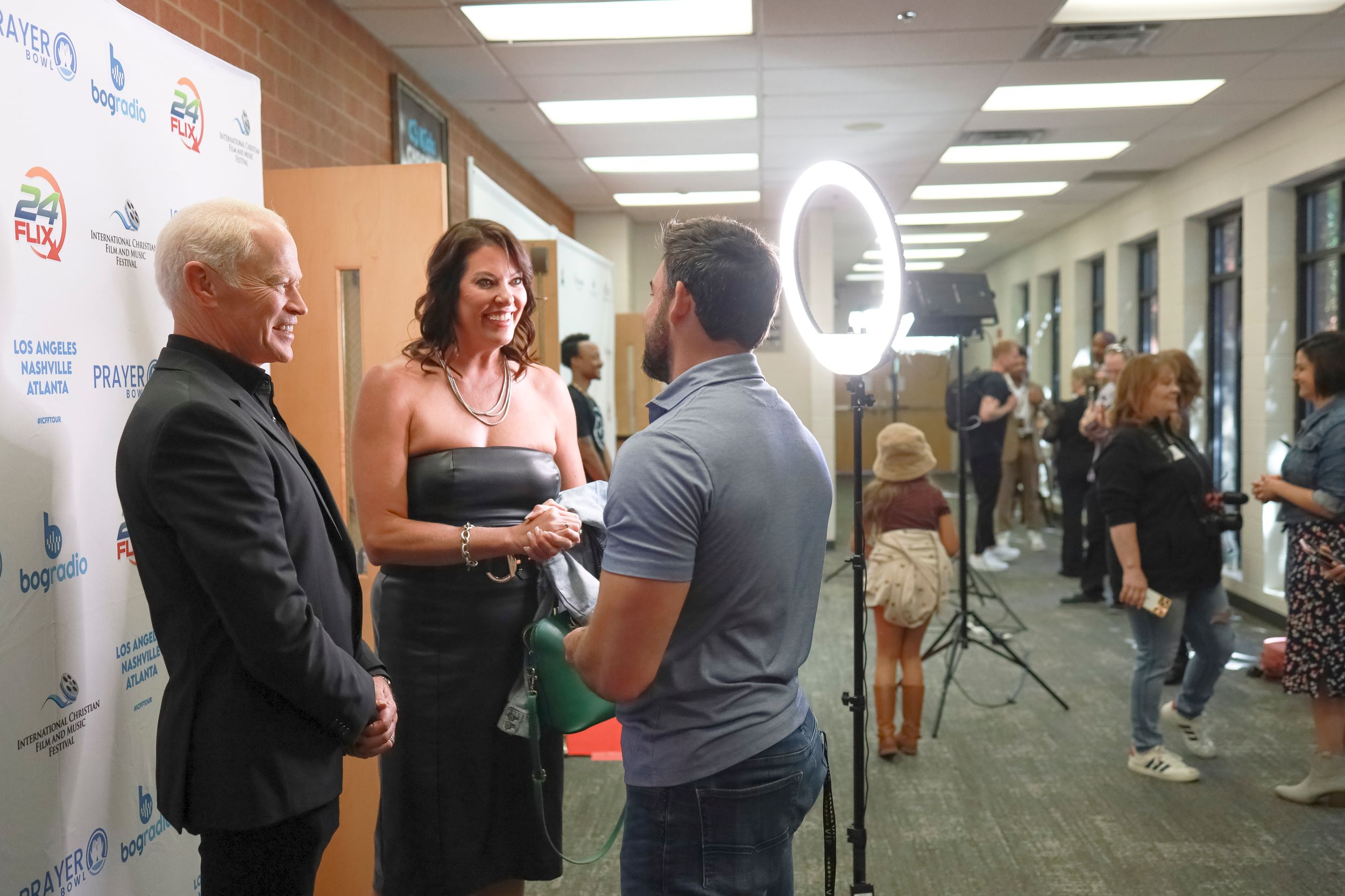 People engaged in conversation during an interview or media event in a brightly lit hallway with camera equipment visible.