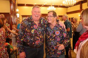 Two people wearing matching colorful floral print shirts smile together at an indoor event with a chandelier.