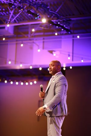 A speaker in a light gray suit holds a microphone while presenting on stage with purple lighting and glowing orbs above.