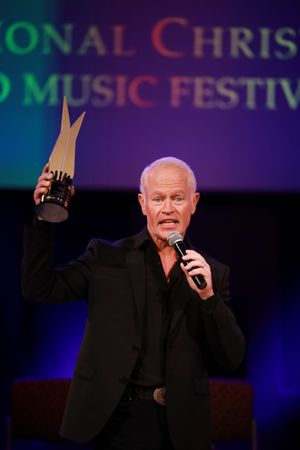 Speaker holds up award trophy while performing on stage at a Christian music festival with colorful backdrop.