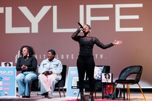 Performer in black attire gestures dramatically while speaking on stage with 'LYLIFE' text displayed behind them.