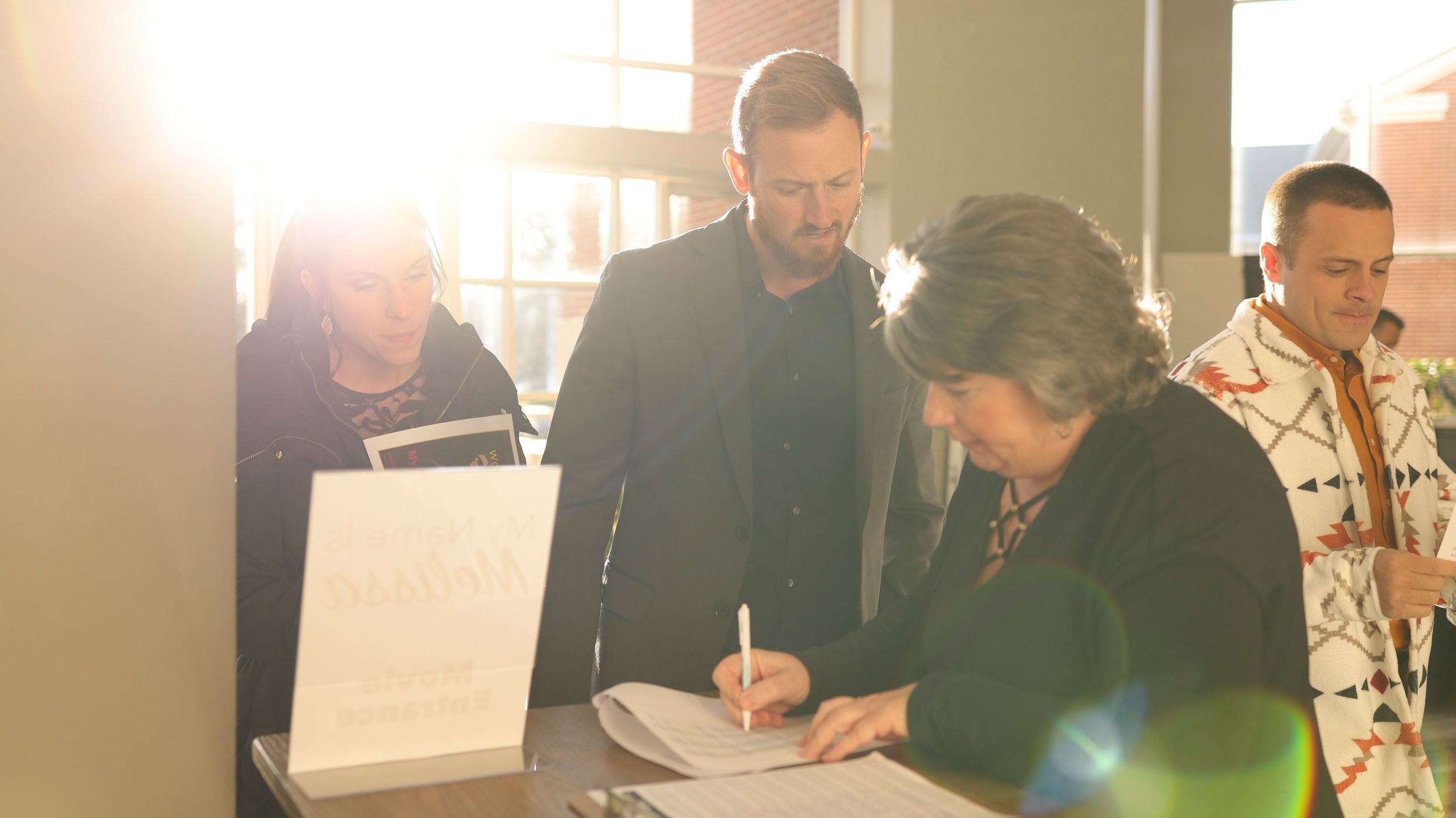 A group of people gathered around a table examining documents together in a sunlit office space.