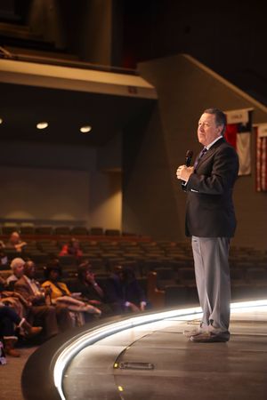A speaker in a dark suit and gray pants stands on a curved illuminated stage addressing an audience in an auditorium.