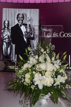 An elegant white floral centerpiece arrangement with lilies and hydrangeas in front of an awards ceremony backdrop.