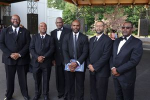 A group of six men in formal black suits and bow ties stand together at an outdoor event.