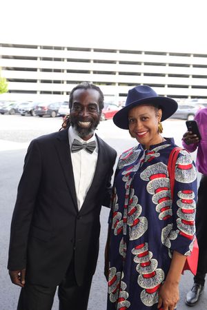 A smiling couple poses together at an event, with one person wearing a black suit and the other in a colorful patterned outfit.