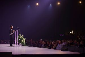 A speaker presents on a dimly lit stage with purple lighting while audience members watch from their seats.