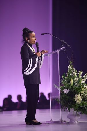 A speaker in a black and white outfit stands at a podium with floral arrangements on a purple-lit stage.
