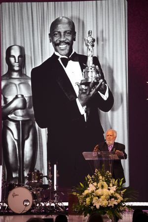 Black and white photos of award ceremony moments are displayed behind a podium with floral arrangements on stage.