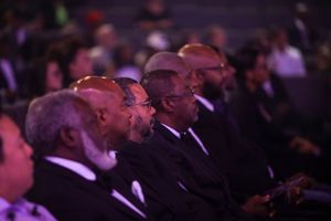 Black-tie audience members seated in a dimly lit theater bathed in purple lighting during a formal event.