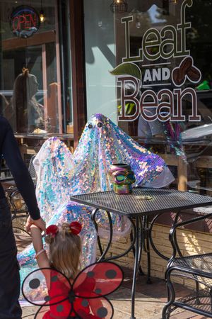 A colorful, sparkly fabric-covered figure sits at an outdoor cafe table in front of a 'Leaf and Bean' storefront.