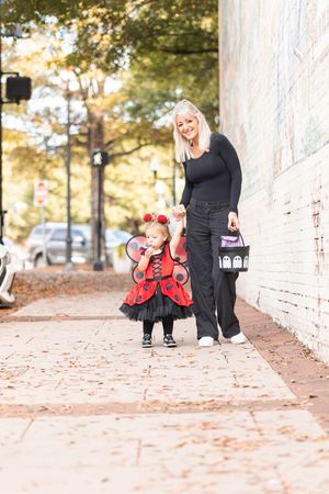 A parent and small child in a ladybug costume walk along a brick sidewalk on an autumn day.