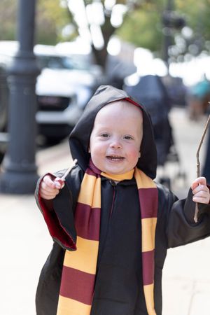 Small child wearing Gryffindor-inspired Harry Potter costume with black robe and maroon and gold striped scarf smiles outdoors.