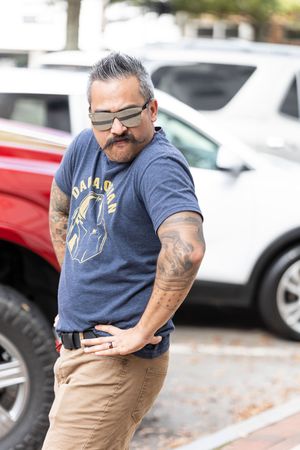 Person wearing glasses and a blue t-shirt with tattoos poses casually in a parking lot with cars in background.
