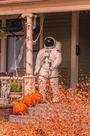 An astronaut suit decoration stands on a porch with pumpkins and scattered autumn leaves for Halloween decor.