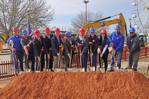 People in construction gear stand in a line with golden shovels during a groundbreaking ceremony on red clay soil.
