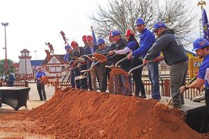 Construction workers observe groundbreaking ceremony with shovels digging into red clay soil at new development site.