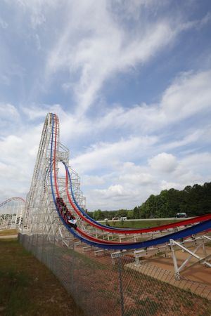 A towering roller coaster track with red and blue rails curves dramatically against a bright cloudy sky.