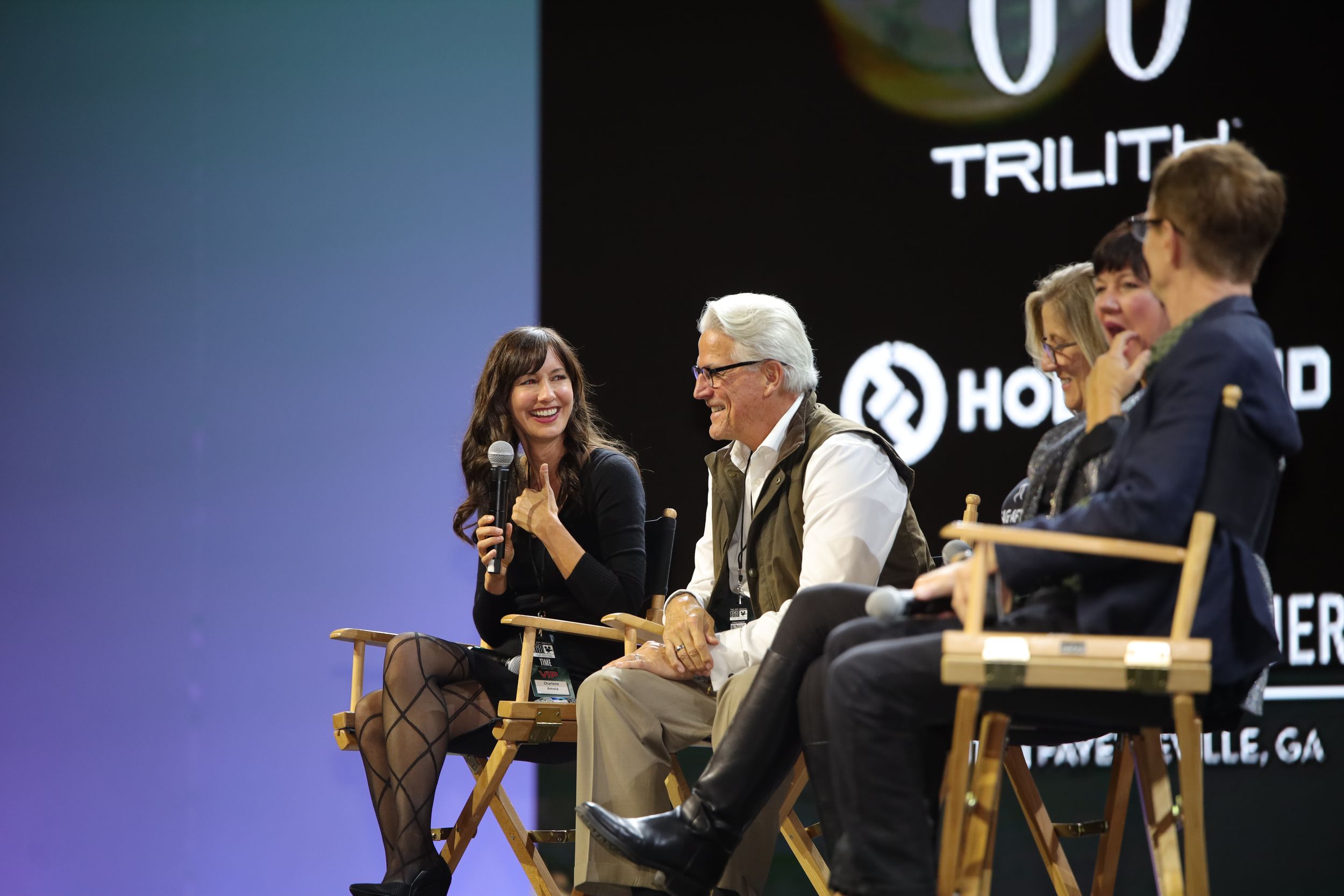 A group of people sit on stage in director's chairs during what appears to be a panel discussion or interview.
