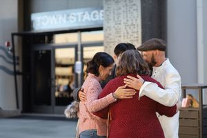 Three friends embrace in a warm group hug outside a storefront entrance with a Stage sign visible above.