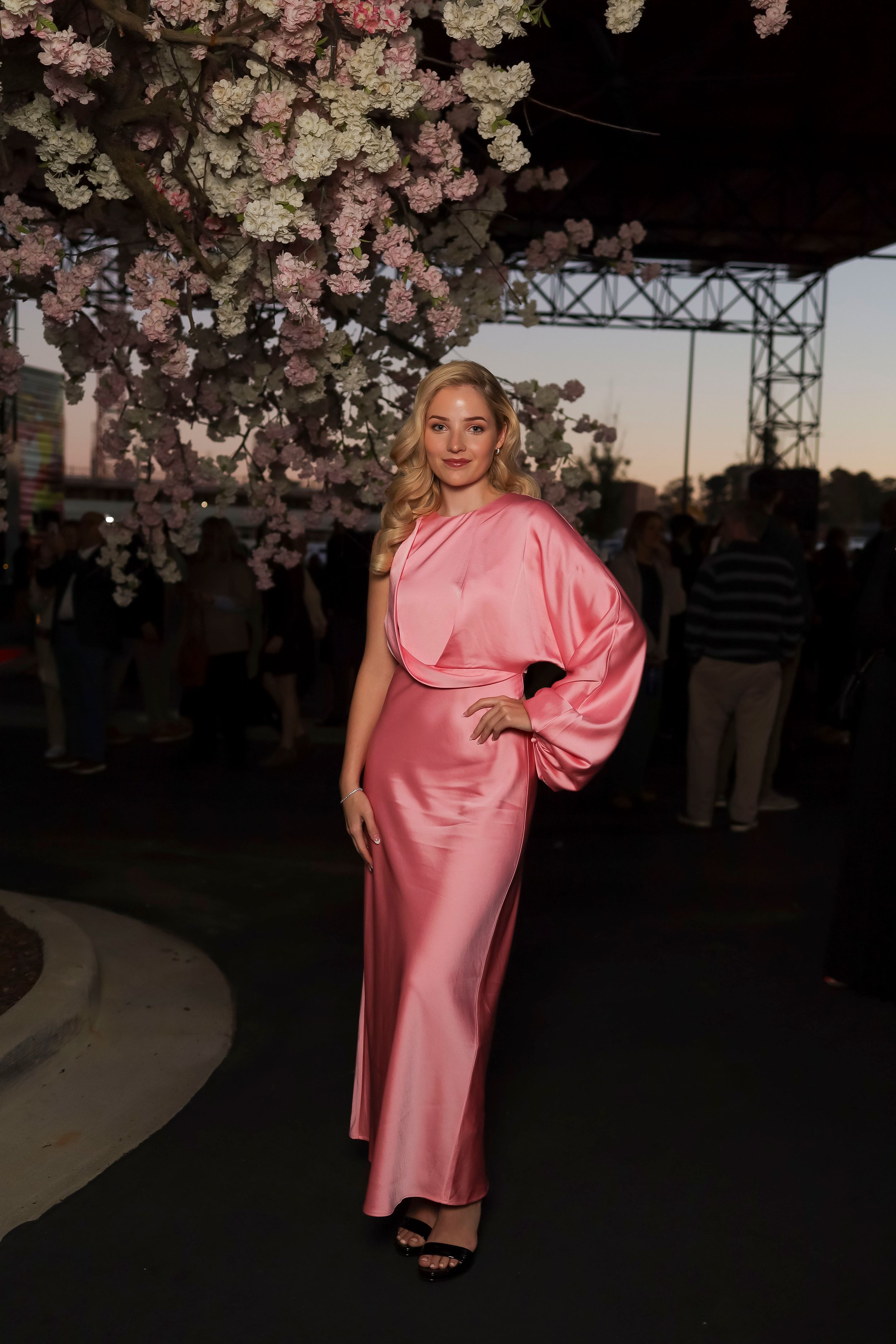 A model poses under cherry blossoms wearing a flowing pink silk dress at dusk.