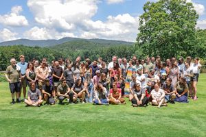 Large group of people gathered on a grassy lawn with green mountains and blue sky in the background.