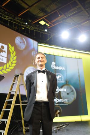 Stage performer in tuxedo stands near wooden ladder against illuminated backdrop at awards ceremony.