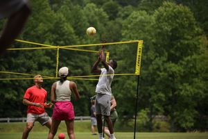 Players compete in a beach volleyball tournament with multiple courts set up in a lush green outdoor setting.