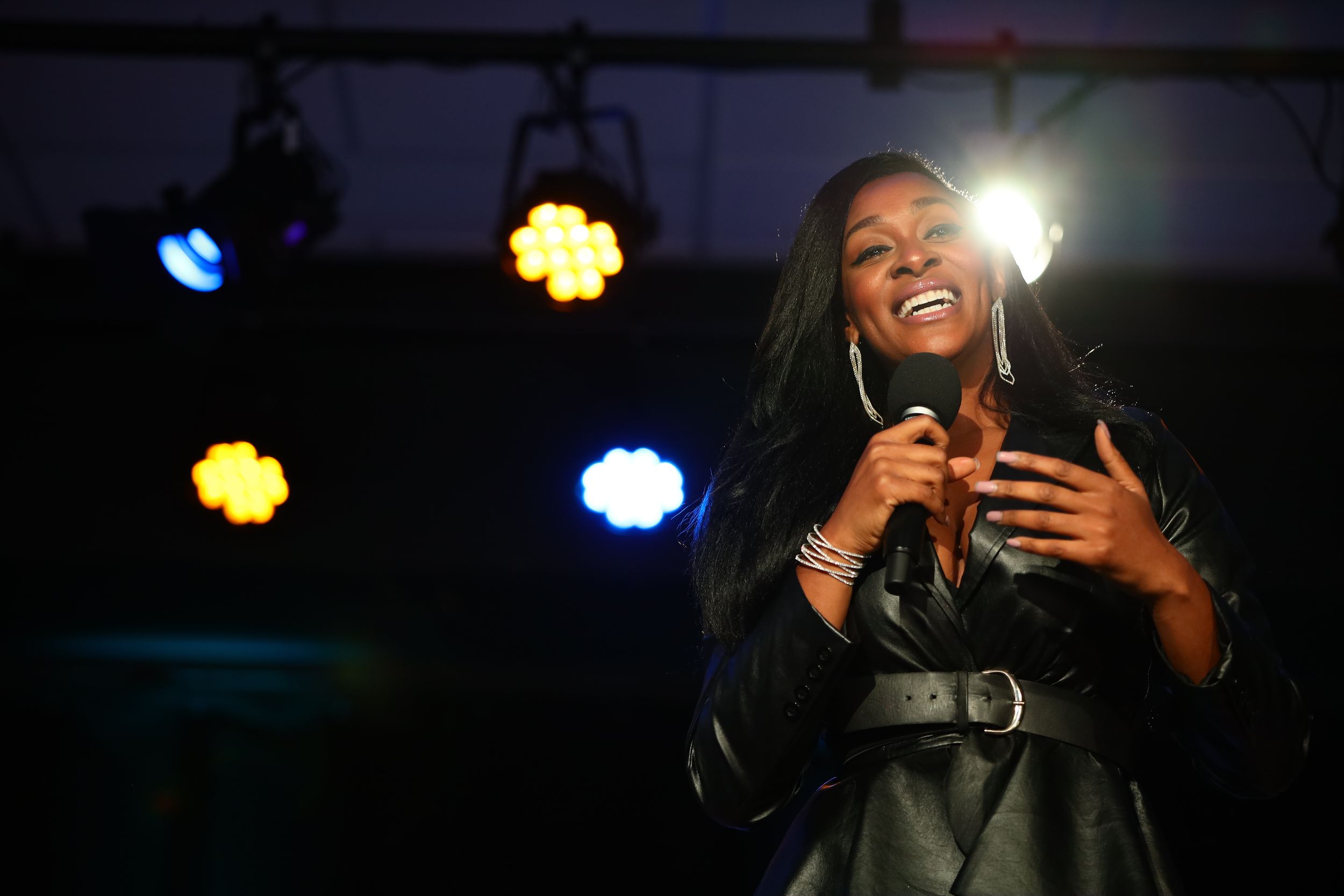 Stage performer in a leather jacket singing with colorful spotlights illuminating the venue behind them.
