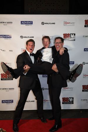 Three performers in black suits strike playful poses on a red carpet at an awards event backdrop.