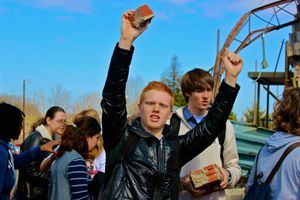 Students celebrating outdoors in a crowd with raised arms against blue sky and metal structure in background.