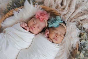 Two sleeping babies in white swaddles lie on fluffy white blanket with pink and blue floral hair accessories.